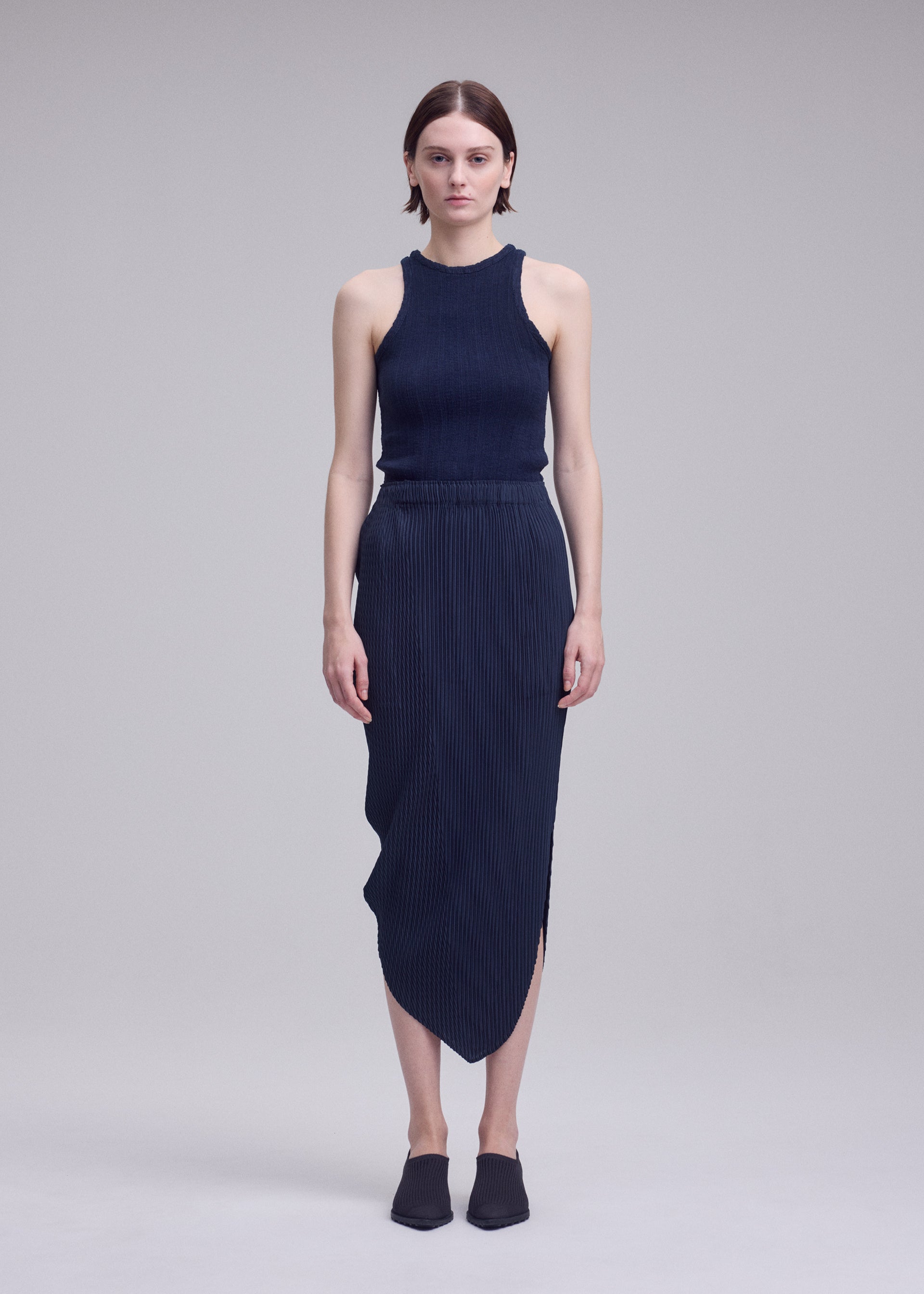 Zoomed view A woman with short hair and a neutral expression stands against a plain background, wearing the ISSEY MIYAKE LEAF PLEATS set—a sleeveless, dark ribbed top paired with a matching leaf silhouette skirt—and black slip-on shoes.
