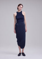 A woman with short brown hair and a relaxed posture stands against a plain background wearing the ISSEY MIYAKE LEAF PLEATS sleeveless navy ribbed top and matching pleated skirt featuring elegant double-pleating detail, paired with black slip-on shoes.