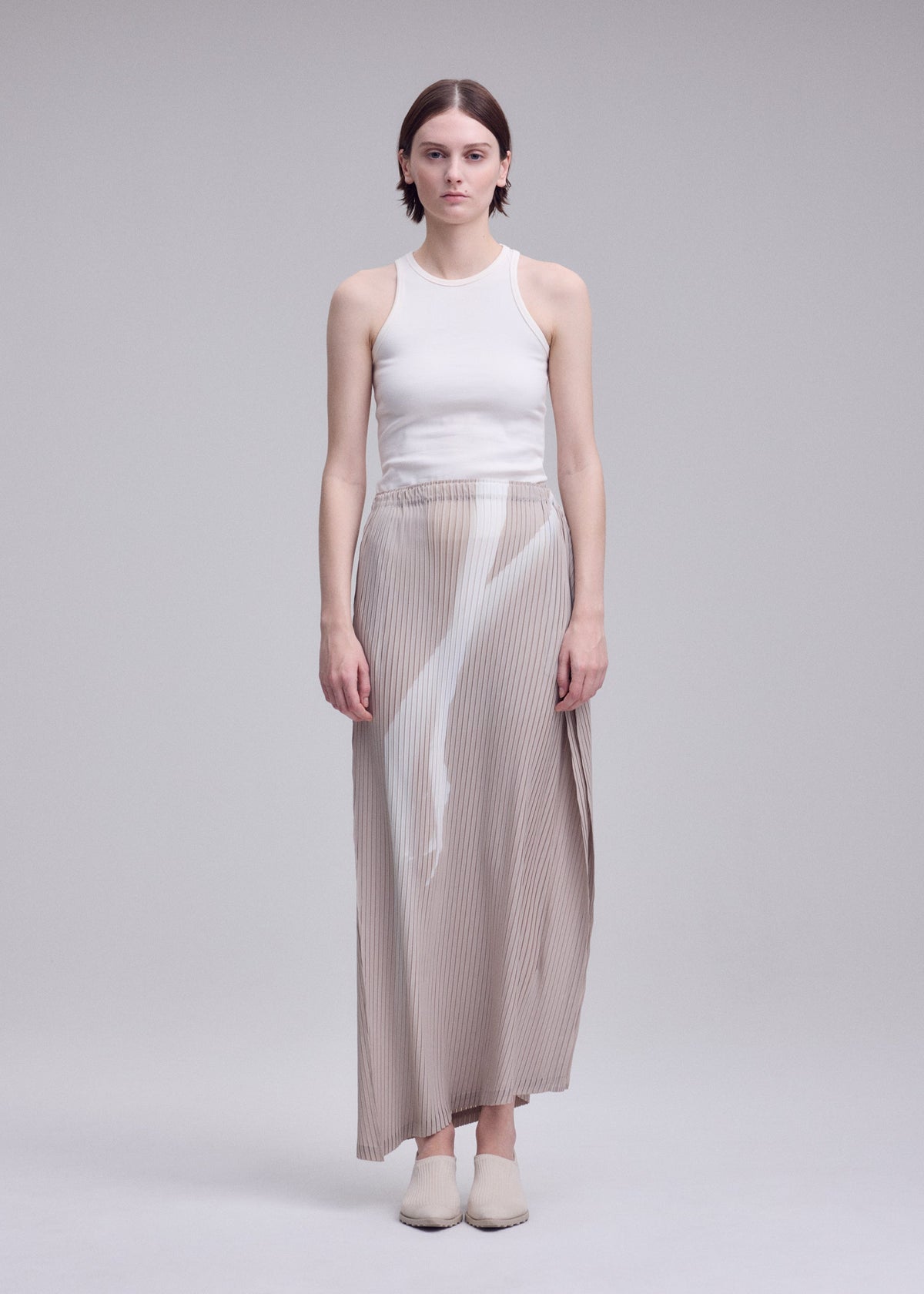 A person stands facing forward in a plain, light gray studio, wearing the HANDS OF LOVER PLEATS skirt by ISSEY MIYAKE—a long, beige polyester crepe de chine piece featuring a white abstract pattern. They are also wearing a white sleeveless top and have short brown hair with a neutral expression. view 5 of 7