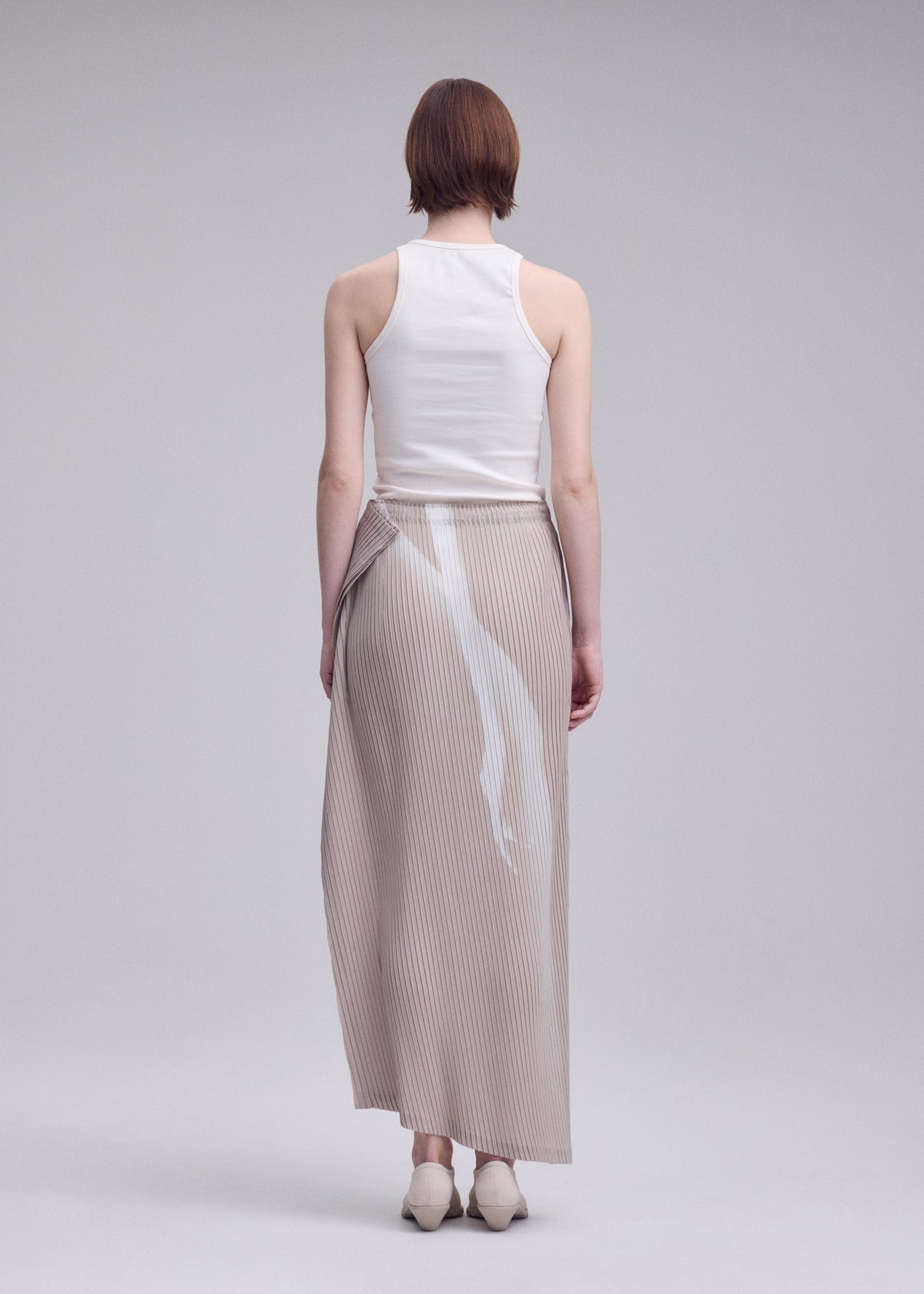 A person with short brown hair, dressed in a white sleeveless top and ISSEY MIYAKE’s HANDS OF LOVER PLEATS—a long beige polyester crepe de chine skirt adorned with original white paint marks—stands facing away from the camera in a plain studio setting. view 6 of 7