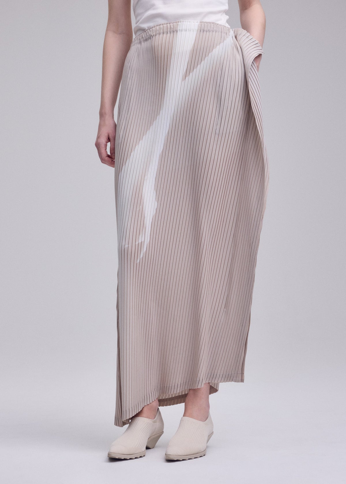 A person wearing a white top, cream-colored shoes, and the ISSEY MIYAKE HANDS OF LOVER PLEATS skirt—a light beige pleated maxi with an original pattern—stands against a plain gray background, their hands relaxed by their sides. view 7 of 7