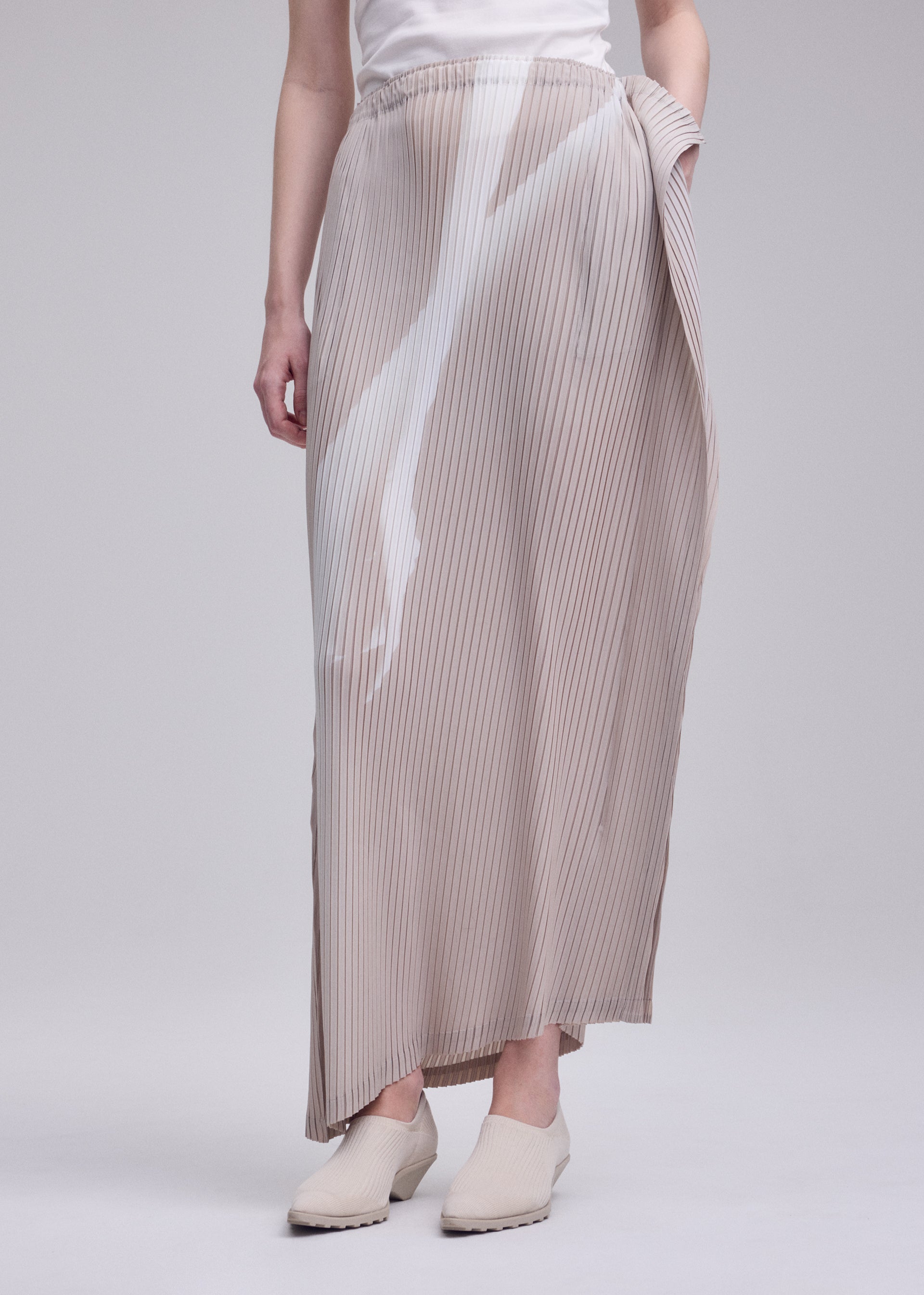 Zoomed view A person wearing a white top, cream-colored shoes, and the ISSEY MIYAKE HANDS OF LOVER PLEATS skirt—a light beige pleated maxi with an original pattern—stands against a plain gray background, their hands relaxed by their sides.