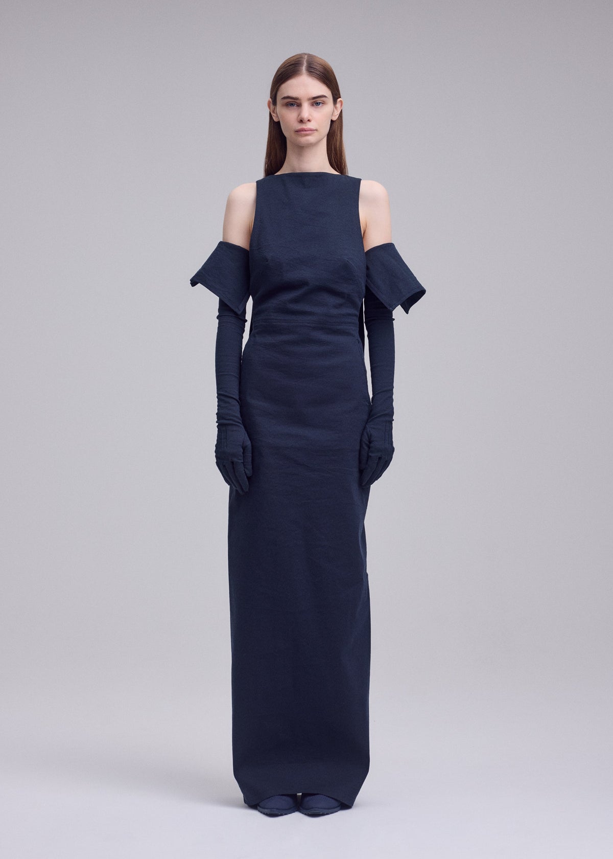 A woman stands facing forward in the ISSEY MIYAKE ALLOW floor-length dark navy minimalist dress with off-the-shoulder sleeves and matching long gloves, her expression neutral against a plain light gray background. view 5 of 8