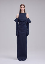 A woman stands facing forward in the ISSEY MIYAKE ALLOW floor-length dark navy minimalist dress with off-the-shoulder sleeves and matching long gloves, her expression neutral against a plain light gray background.