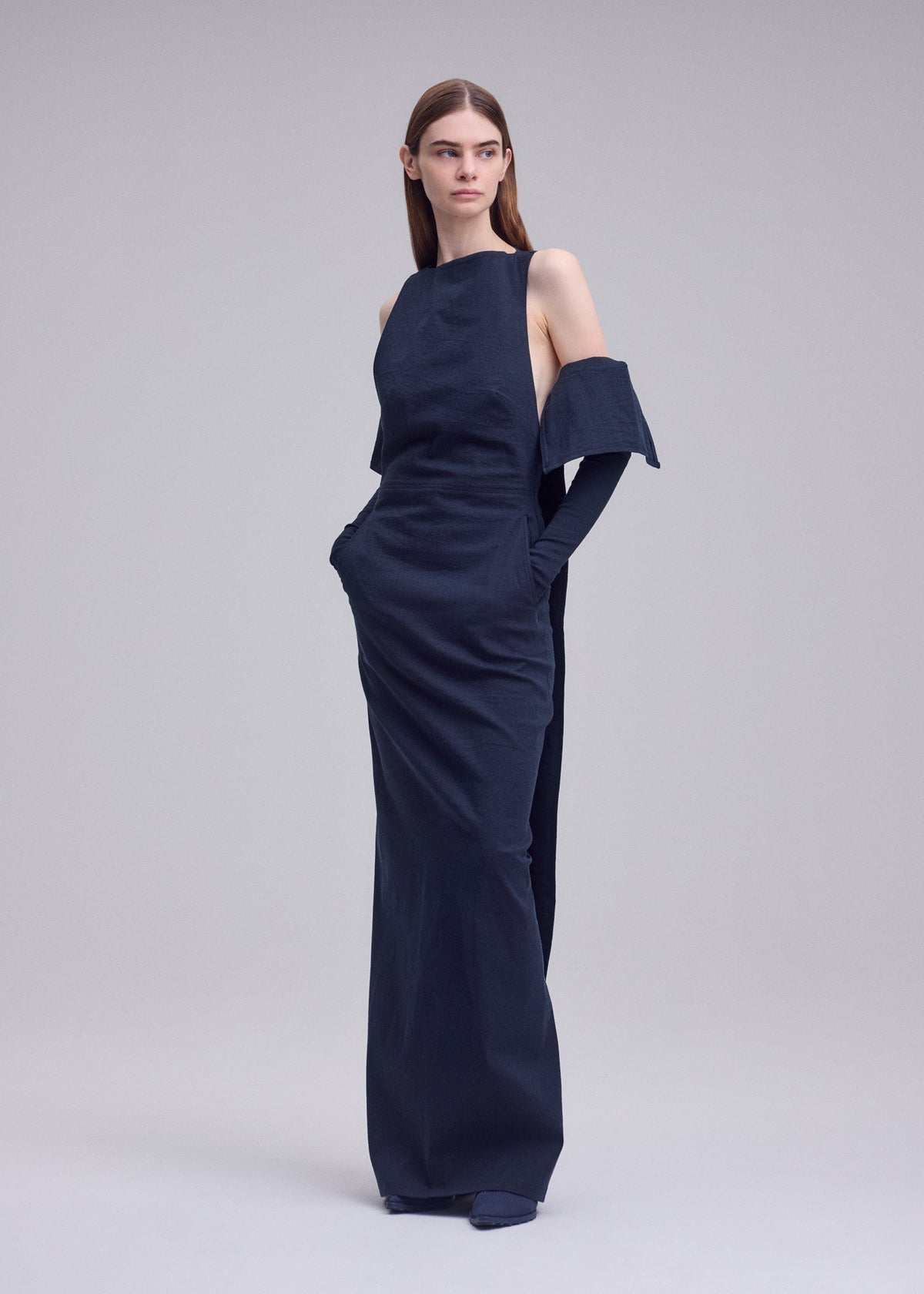 A woman with straight brown hair stands in a studio wearing ALLOW by ISSEY MIYAKE, a long, dark navy-blue washi paper-blend sleeveless dress with pockets and matching gloves, featuring a minimalist, structured design. view 6 of 8