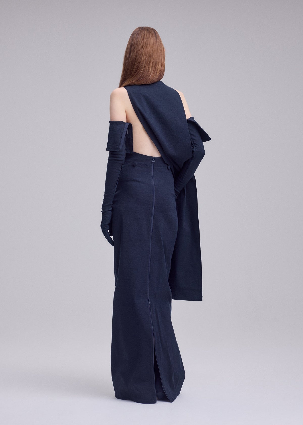 A person with long, straight hair faces away from the camera in ISSEY MIYAKE's ALLOW navy minimalist dress, featuring gloves and draped washi-blend fabric that adds sophistication against a plain, light-hued backdrop. view 7 of 8
