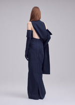 A person with long, straight hair faces away from the camera in ISSEY MIYAKE's ALLOW navy minimalist dress, featuring gloves and draped washi-blend fabric that adds sophistication against a plain, light-hued backdrop.