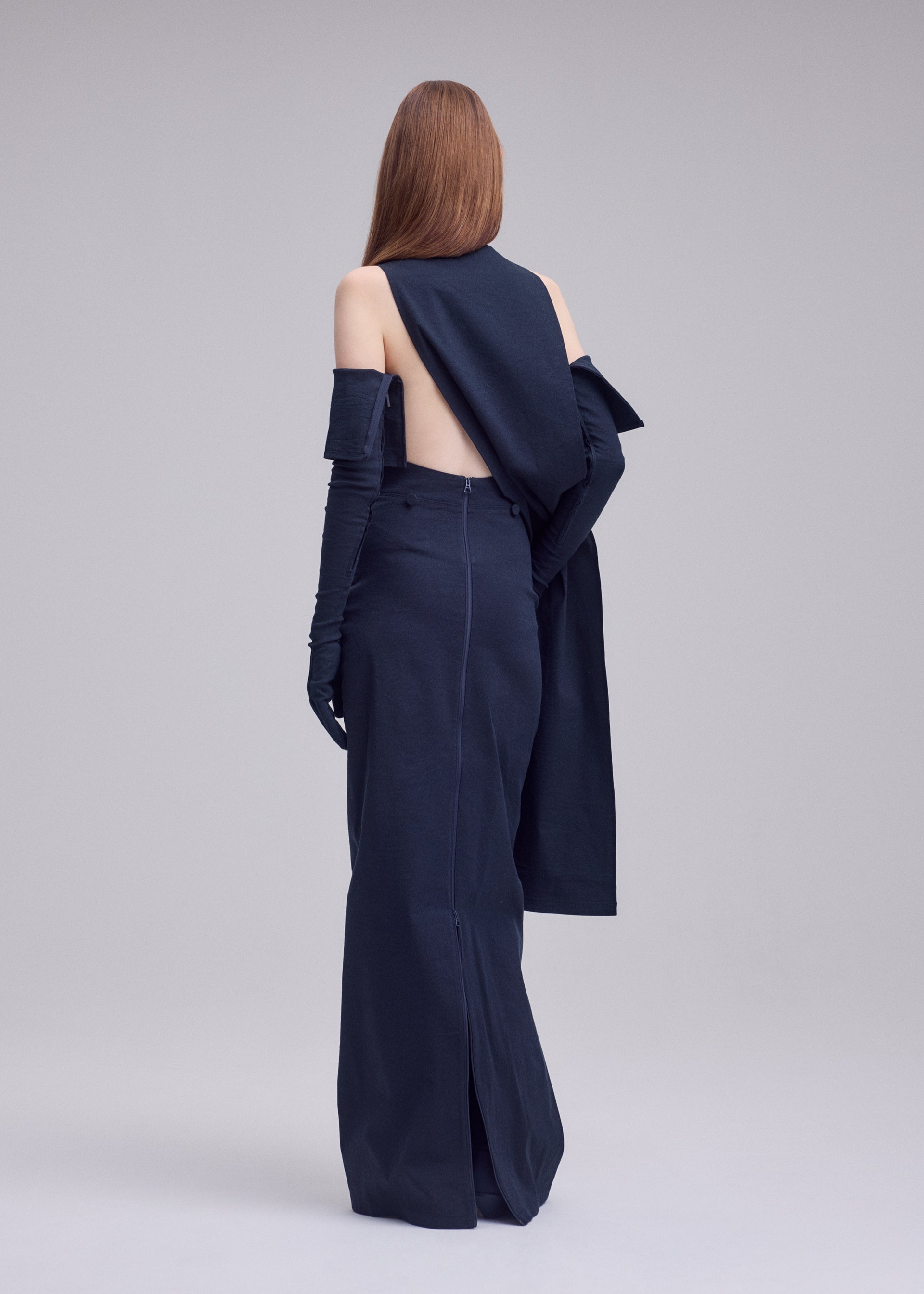 Zoomed view A person with long, straight hair faces away from the camera in ISSEY MIYAKE's ALLOW navy minimalist dress, featuring gloves and draped washi-blend fabric that adds sophistication against a plain, light-hued backdrop.