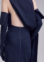 A person wears the ALLOW dress by ISSEY MIYAKE—a navy blue, backless minimalist piece with sleek lines and long matching gloves. The photo, taken from behind, highlights its open back and skirt zipper for an understated look.