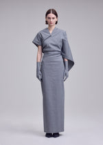 A woman poses against a gray backdrop in ISSEY MIYAKE’s ALLOW dress—a long, fitted minimalist design with an asymmetric sleeve, matching gray gloves, dark shoes, and a subtle stone-like texture. Her expression is neutral.