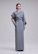 A person with short brown hair and a neutral expression stands against a plain background, wearing the ALLOW long gray minimalist dress by ISSEY MIYAKE, featuring a stone-like texture, asymmetrical sleeves, and matching gloves.