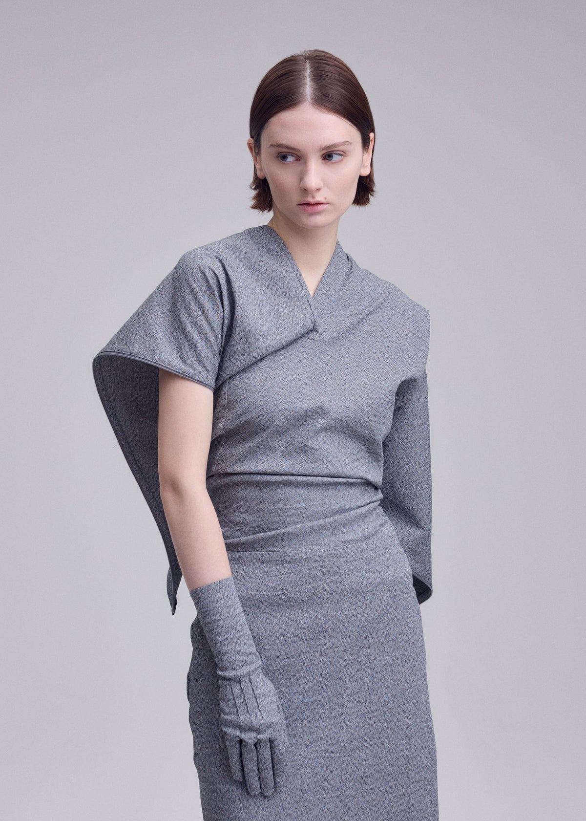A woman with short brown hair wears the grey ALLOW dress by ISSEY MIYAKE, featuring an asymmetrical sleeve, matching glove, and stone-like texture. She stands against a plain light background, looking to the side with a neutral expression. view 7 of 8