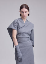 A woman with short brown hair wears the grey ALLOW dress by ISSEY MIYAKE, featuring an asymmetrical sleeve, matching glove, and stone-like texture. She stands against a plain light background, looking to the side with a neutral expression.