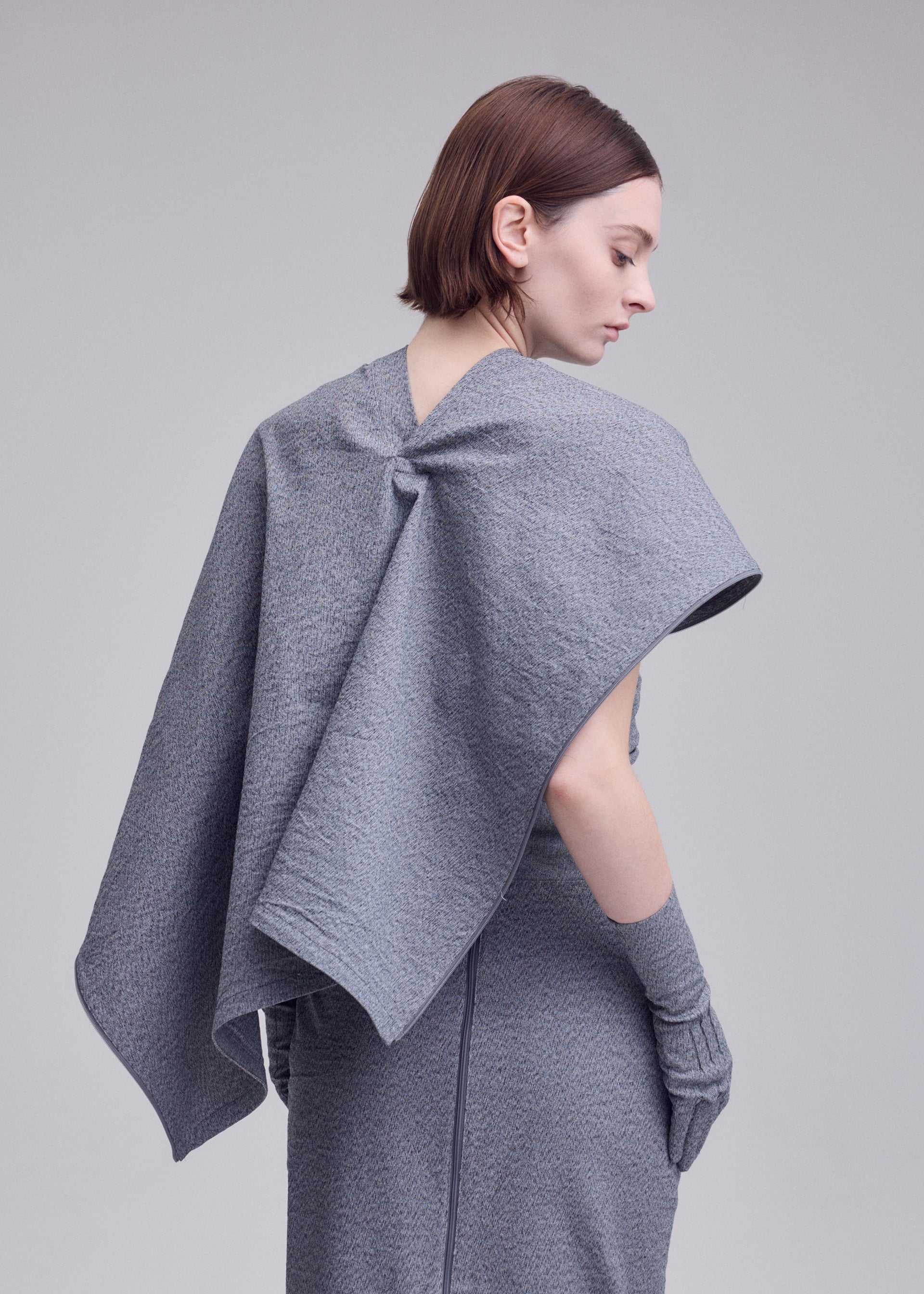 Zoomed view A woman with short brown hair stands sideways, modeling the ALLOW dress by ISSEY MIYAKE—crafted from a washi paper blend with a stone-like texture, styled with a cape and matching fingerless gloves against a plain background.
