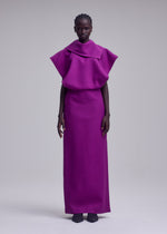A person stands against a plain background wearing the CALLIGRAPH, a floor-length, bright magenta premium wool dress by ISSEY MIYAKE, featuring wide structured shoulders and a high collar. Paired with black shoes, the look recalls ISSEY MIYAKE’s signature sculptural silhouettes.