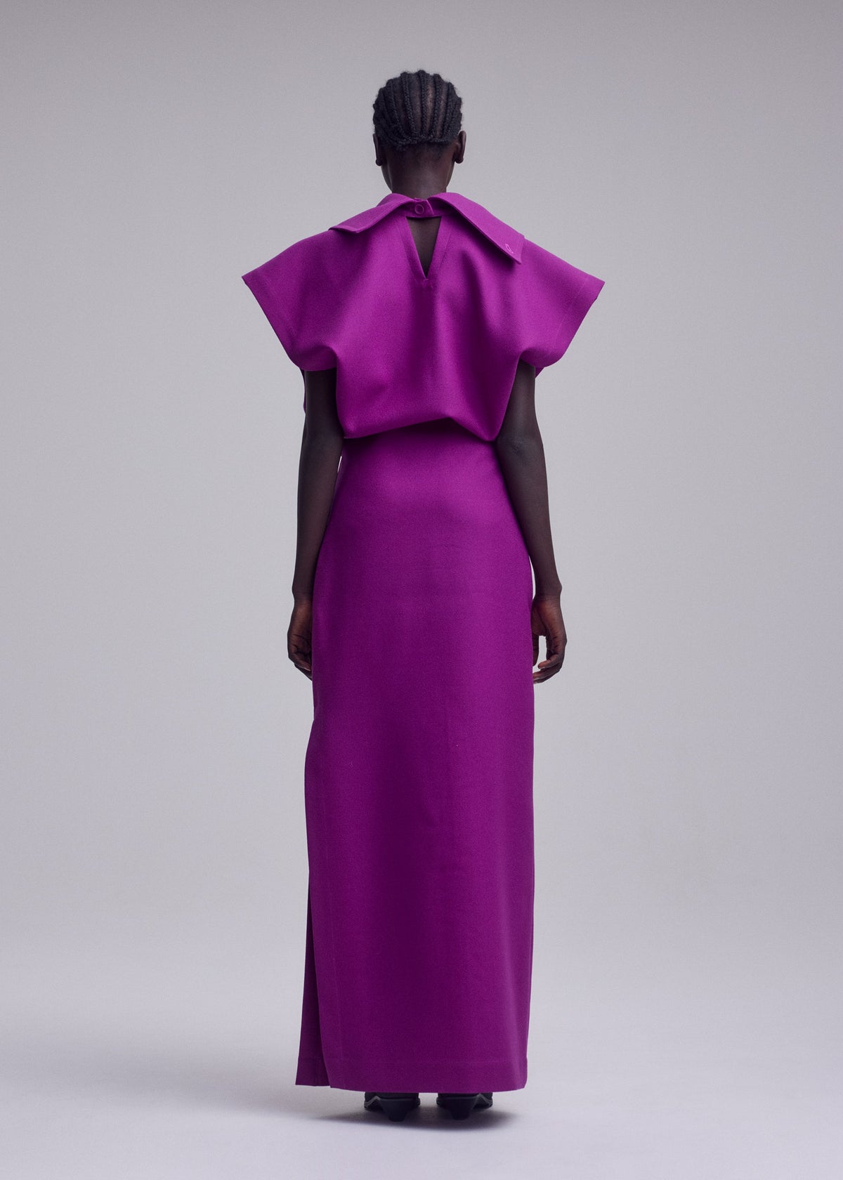 A person stands facing away, wearing the ISSEY MIYAKE CALLIGRAPH dress—a vibrant purple, floor-length piece crafted from premium wool with wide sleeves and a small back keyhole detail. The background is plain and light-colored. view 5 of 7