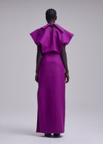 A person stands facing away, wearing the ISSEY MIYAKE CALLIGRAPH dress—a vibrant purple, floor-length piece crafted from premium wool with wide sleeves and a small back keyhole detail. The background is plain and light-colored.