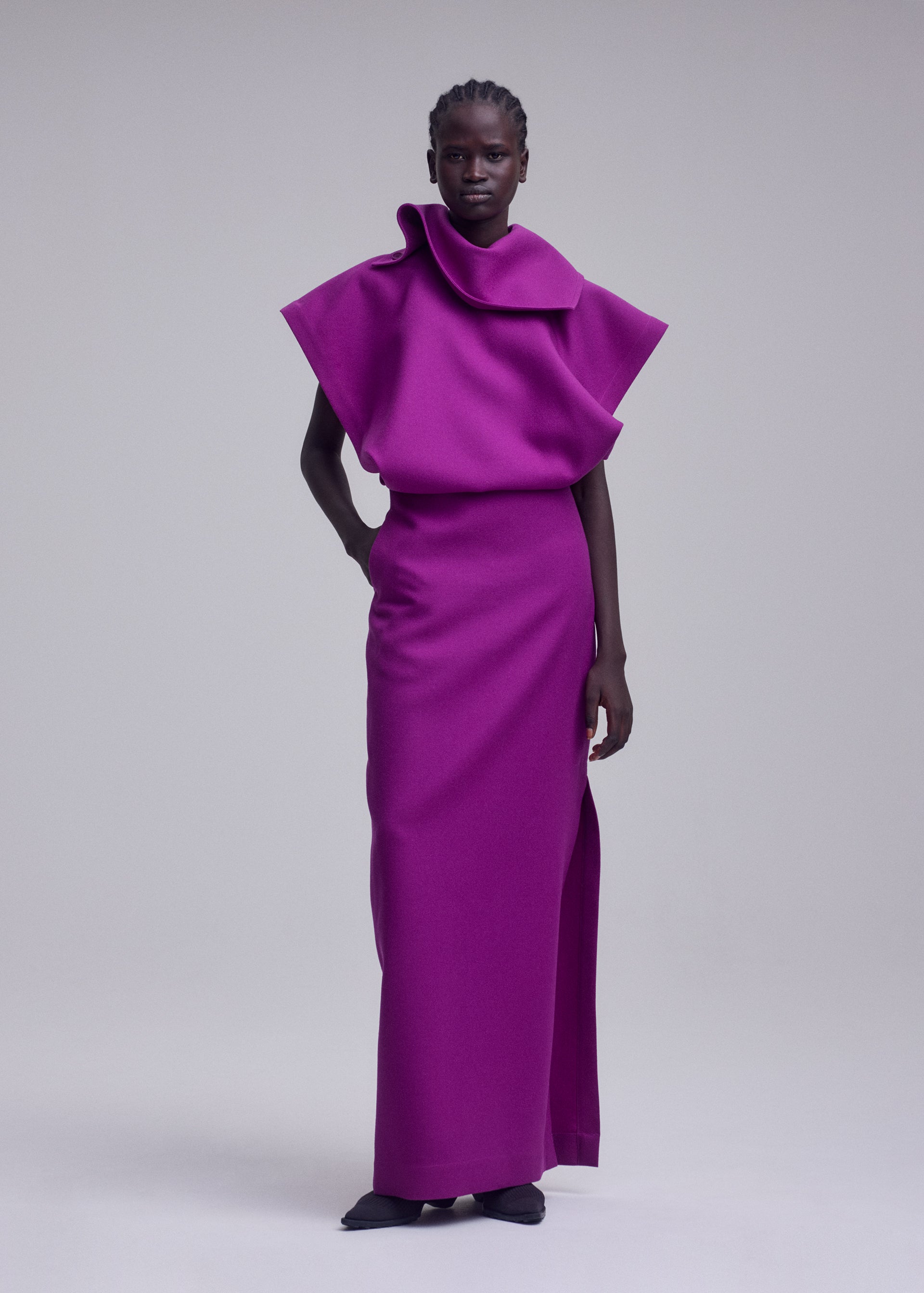 Zoomed view A person stands wearing the CALLIGRAPH ensemble by ISSEY MIYAKE—a vibrant magenta, sculptural top with wide shoulders paired with a matching long skirt, both crafted from premium wool dress material. Evoking ISSEY MIYAKE’s signature bold and minimalist aesthetic, the outfit features striking lines and effortless elegance. The background is plain and light-colored.