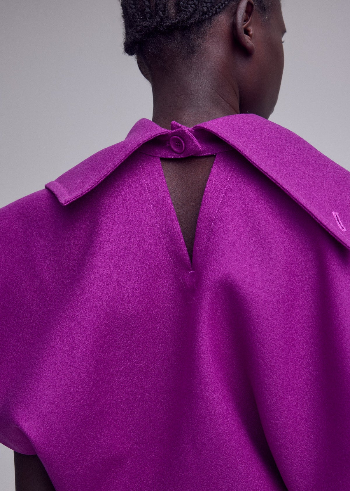 A person wearing the CALLIGRAPH dress by ISSEY MIYAKE—a vibrant purple premium wool piece featuring a V-shaped back opening and button closure, crafted from the brand’s signature nashiji pebbled fabric—is photographed from behind against a plain background. view 7 of 7