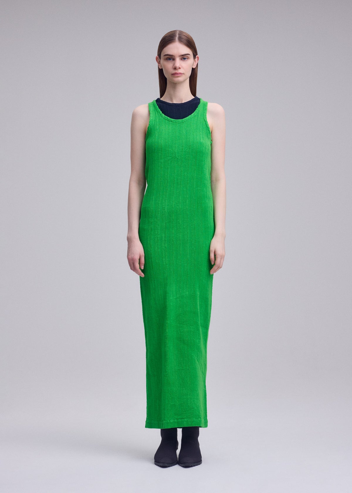 A person faces forward wearing ISSEY MIYAKE's RILL, an ankle-length sleeveless bright green pleated dress over a black top with dark shoes. The subtly textured garment stands out against a plain light gray background. view 5 of 6