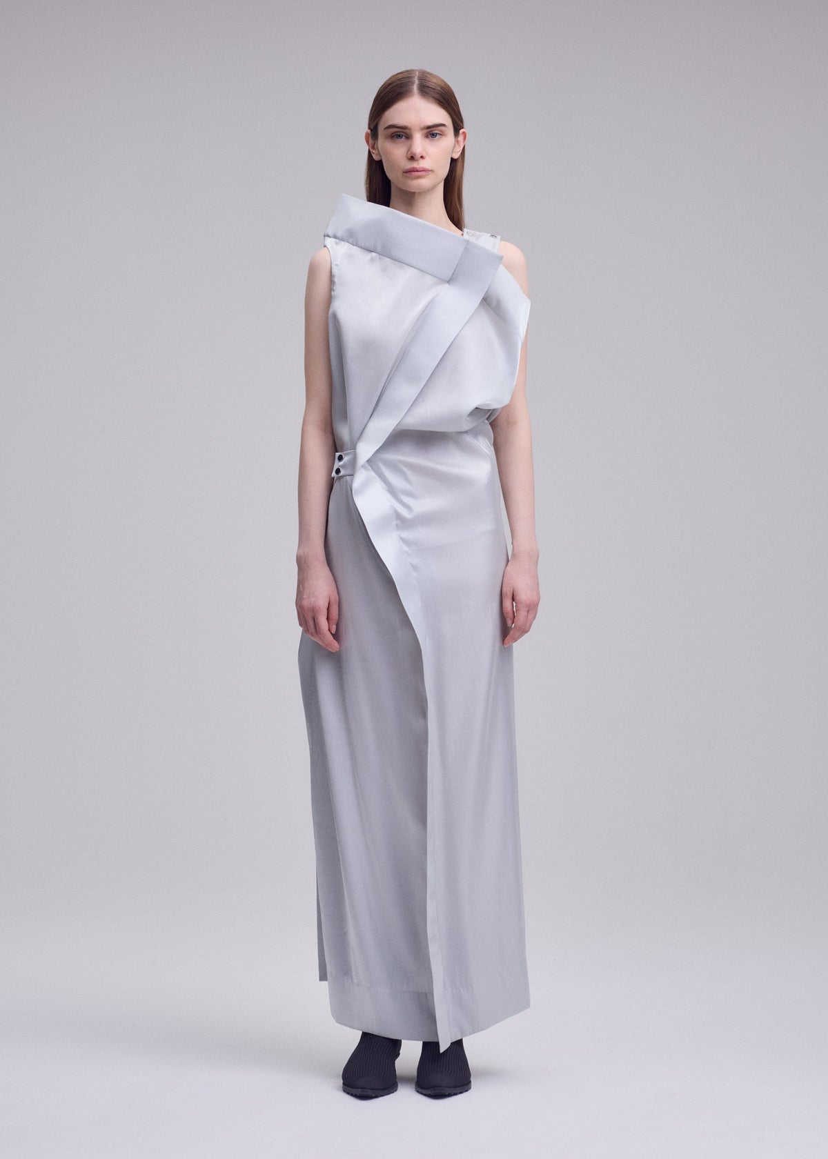 A woman faces forward against a plain light gray background, wearing the MERCURY dress by ISSEY MIYAKE—a sleek silver-gray metallic design with asymmetrical draping and button details—paired with black socks. view 5 of 7