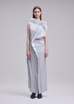 A woman faces forward against a plain light gray background, wearing the MERCURY dress by ISSEY MIYAKE—a sleek silver-gray metallic design with asymmetrical draping and button details—paired with black socks.