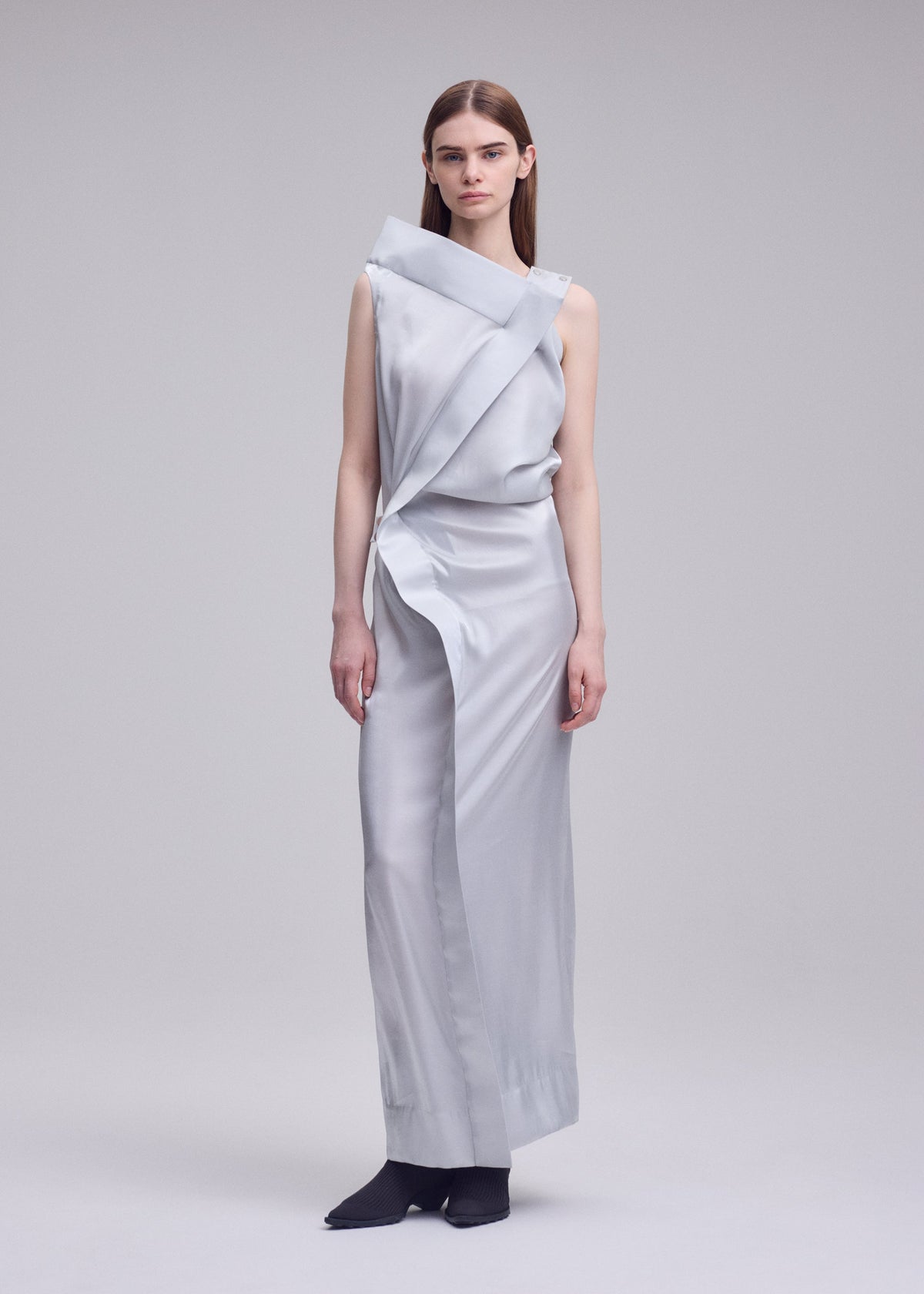 A woman with straight brown hair and a neutral expression stands barefoot against a plain background, wearing the ISSEY MIYAKE MERCURY dress—a sleek metallic piece featuring an asymmetrical neckline and draped design. view 6 of 7