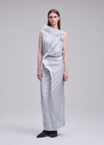 A woman with straight brown hair and a neutral expression stands barefoot against a plain background, wearing the ISSEY MIYAKE MERCURY dress—a sleek metallic piece featuring an asymmetrical neckline and draped design.