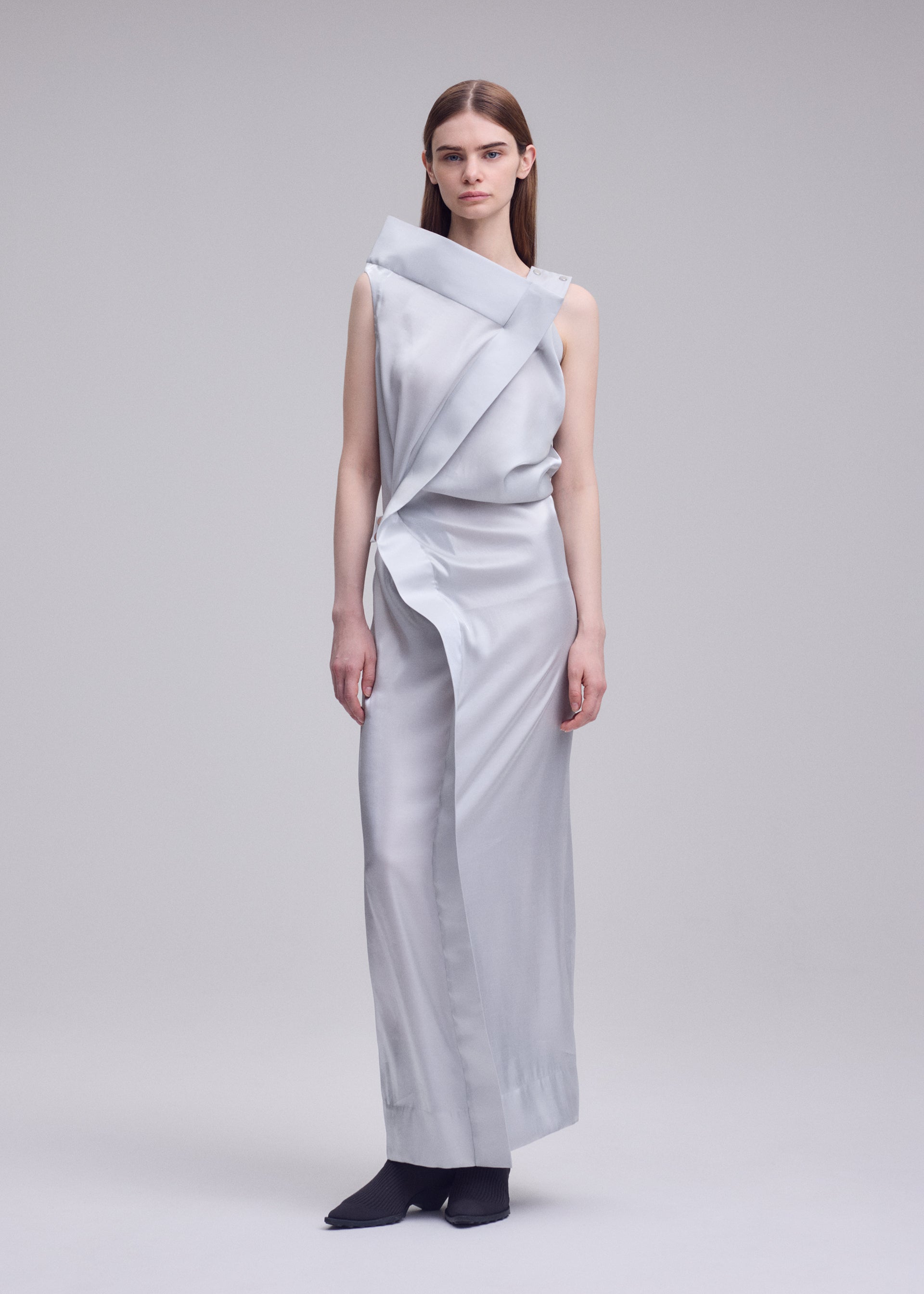 Zoomed view A woman with straight brown hair and a neutral expression stands barefoot against a plain background, wearing the ISSEY MIYAKE MERCURY dress—a sleek metallic piece featuring an asymmetrical neckline and draped design.