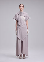 A woman stands against a plain background, wearing the HANDS OF LOVER dress by ISSEY MIYAKE—a long, light gray poplin piece featuring layered, geometric panels that form an abstract pattern. She completes the look with white gloves and shoes, her hair tied back as she faces forward with a neutral expression.