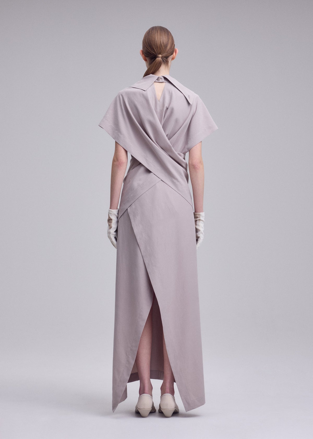 A person stands facing away in a minimal, studio-lit setting, wearing the ISSEY MIYAKE HANDS OF LOVER long light gray poplin dress with layered fabric details, paired with white gloves and cream shoes. view 4 of 6