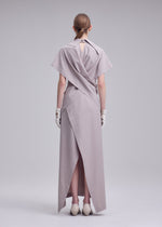 A person stands facing away in a minimal, studio-lit setting, wearing the ISSEY MIYAKE HANDS OF LOVER long light gray poplin dress with layered fabric details, paired with white gloves and cream shoes.