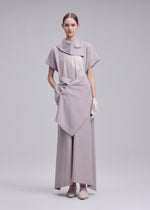 A woman stands against a plain backdrop wearing HANDS OF LOVER, an avant-garde light gray poplin dress by ISSEY MIYAKE, featuring layered asymmetric panels with subtle abstract patterns. She pairs the look with white gloves and matching shoes, her hair pulled back.