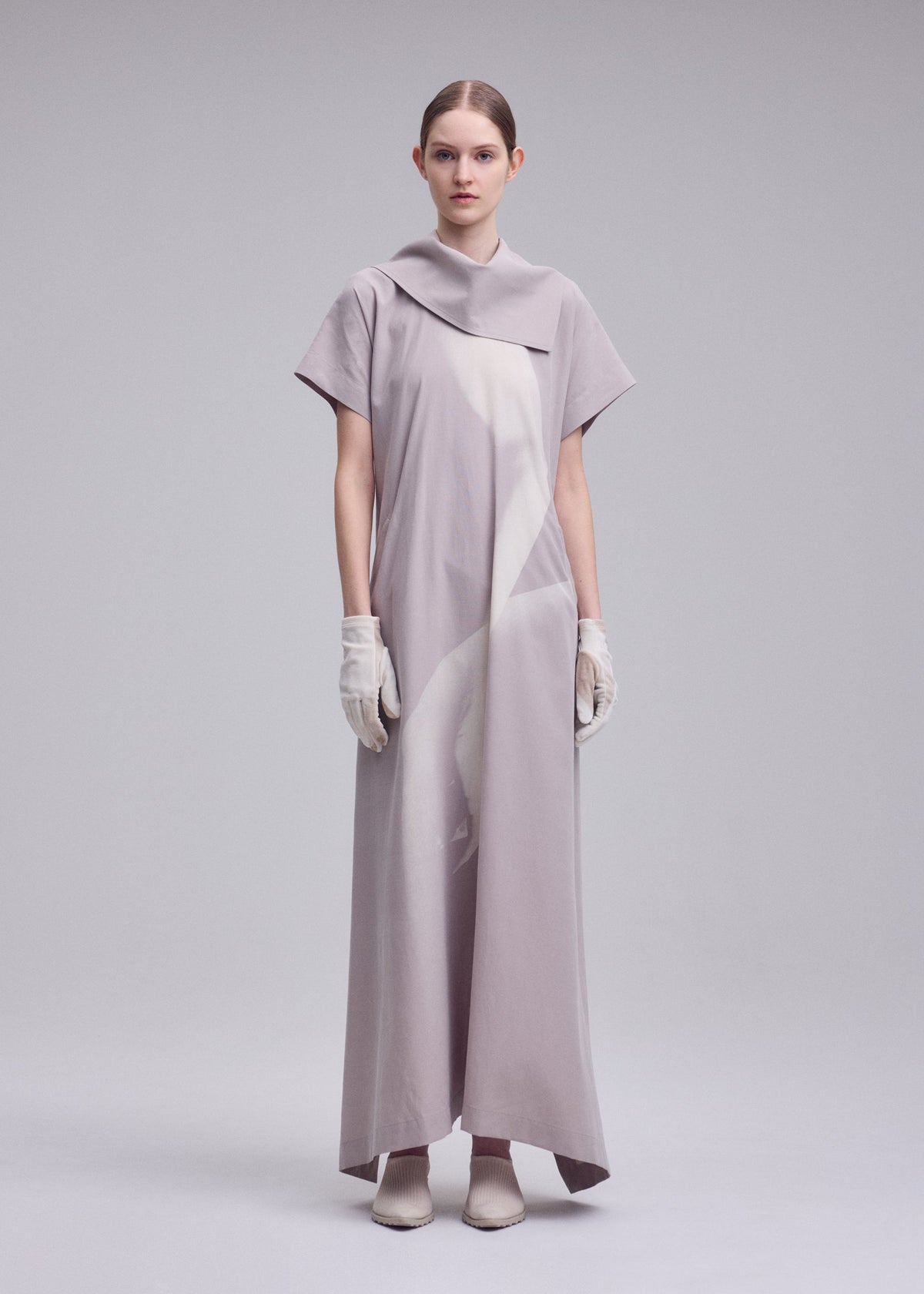 A person stands against a light gray background, wearing the ISSEY MIYAKE HANDS OF LOVER dress—a long, loose-fitting piece featuring a monochrome pattern, short sleeves, and an asymmetrical collar. Crafted from soft poplin fabric, the dress is styled with white gloves and pointed shoes, while their hair is pulled back for a refined look. view 6 of 6