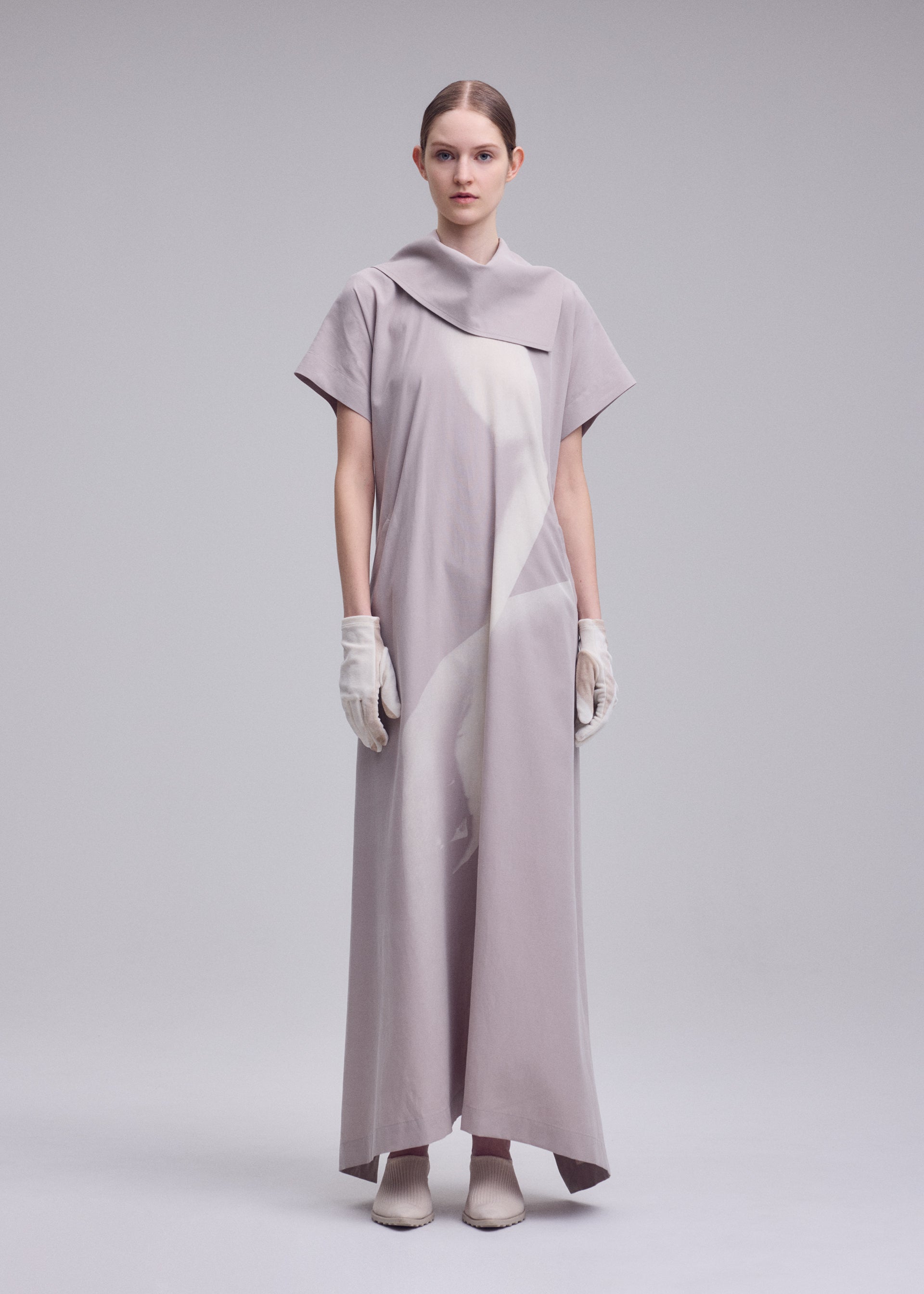 Zoomed view A person stands against a light gray background, wearing the ISSEY MIYAKE HANDS OF LOVER dress—a long, loose-fitting piece featuring a monochrome pattern, short sleeves, and an asymmetrical collar. Crafted from soft poplin fabric, the dress is styled with white gloves and pointed shoes, while their hair is pulled back for a refined look.