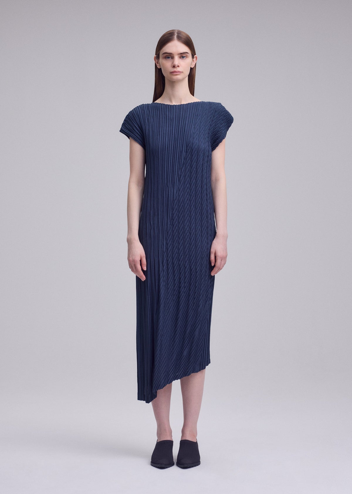 A woman faces forward in ISSEY MIYAKE's UNFURLING PLEATS—a dark blue, short-sleeved, round-neck dress with diagonal pleats and an asymmetrical hem. She has straight brown hair, black slip-ons, and stands against a plain light gray background. view 5 of 6