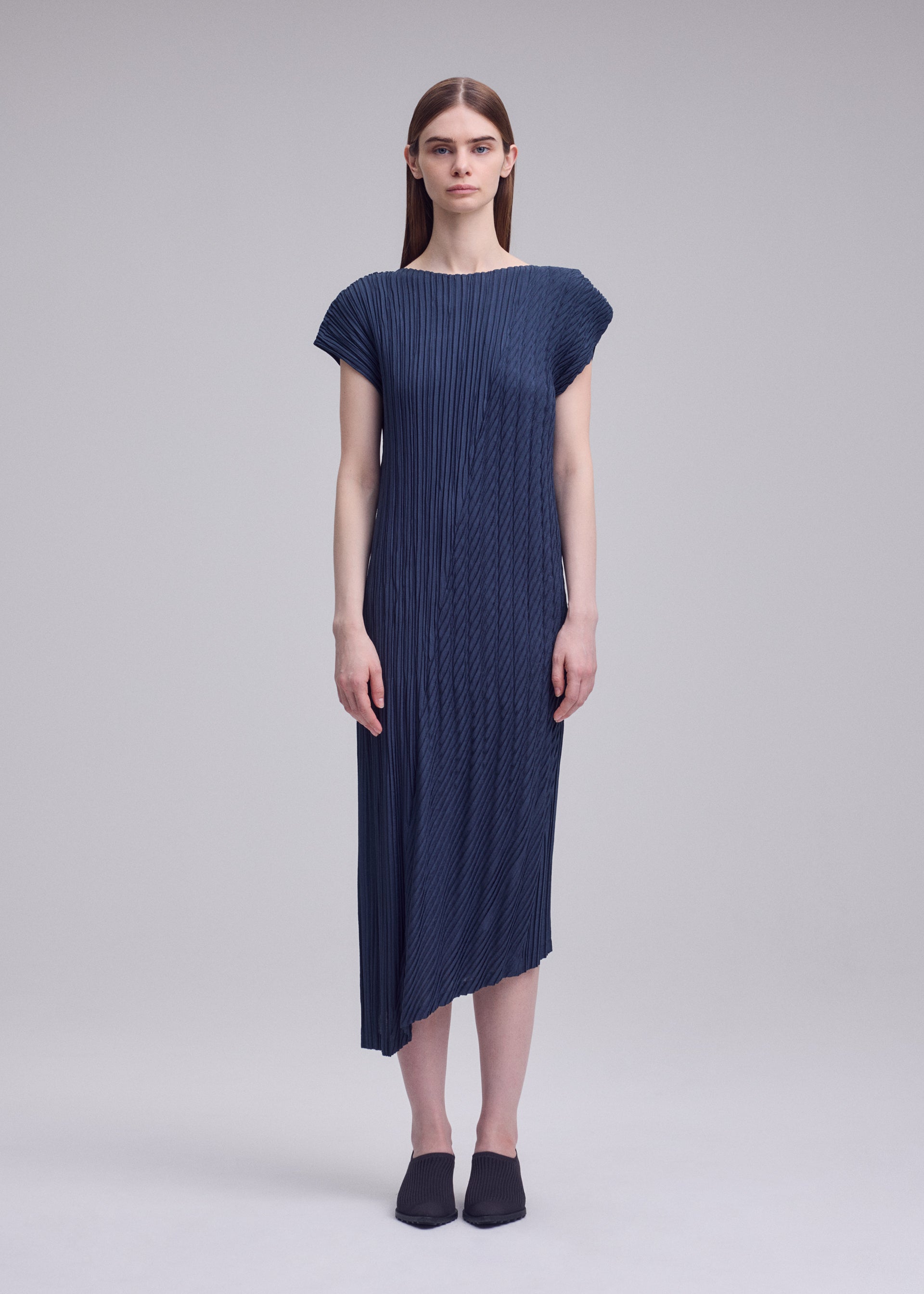 Zoomed view A woman faces forward in ISSEY MIYAKE's UNFURLING PLEATS—a dark blue, short-sleeved, round-neck dress with diagonal pleats and an asymmetrical hem. She has straight brown hair, black slip-ons, and stands against a plain light gray background.