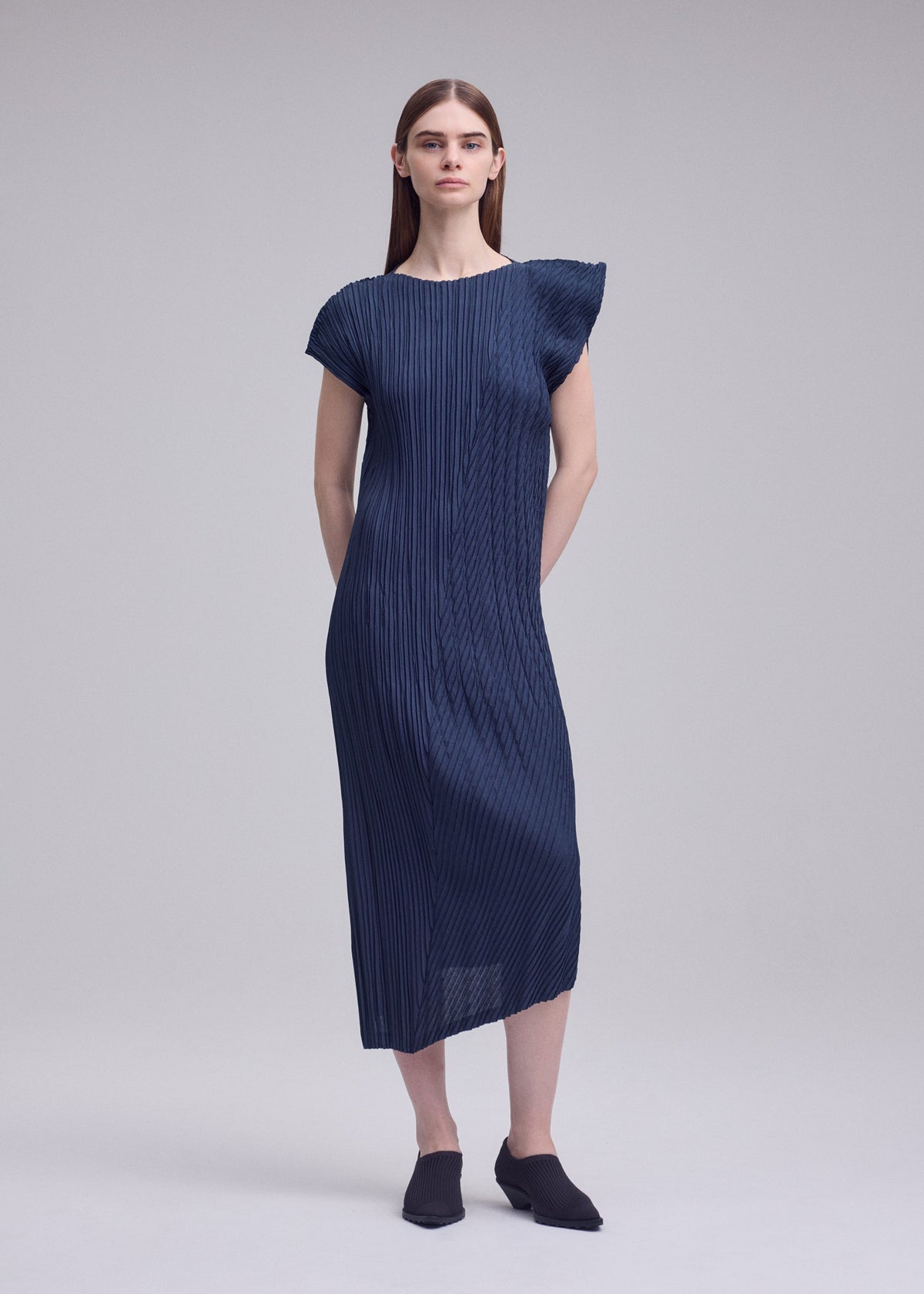 A woman with straight brown hair and a neutral expression wears the ISSEY MIYAKE UNFURLING PLEATS navy blue short-sleeve midi dress with Keshi and diagonal pleating, asymmetrical details, and black slip-on shoes against a plain background. view 6 of 6
