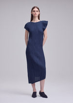 A woman with straight brown hair and a neutral expression wears the ISSEY MIYAKE UNFURLING PLEATS navy blue short-sleeve midi dress with Keshi and diagonal pleating, asymmetrical details, and black slip-on shoes against a plain background.