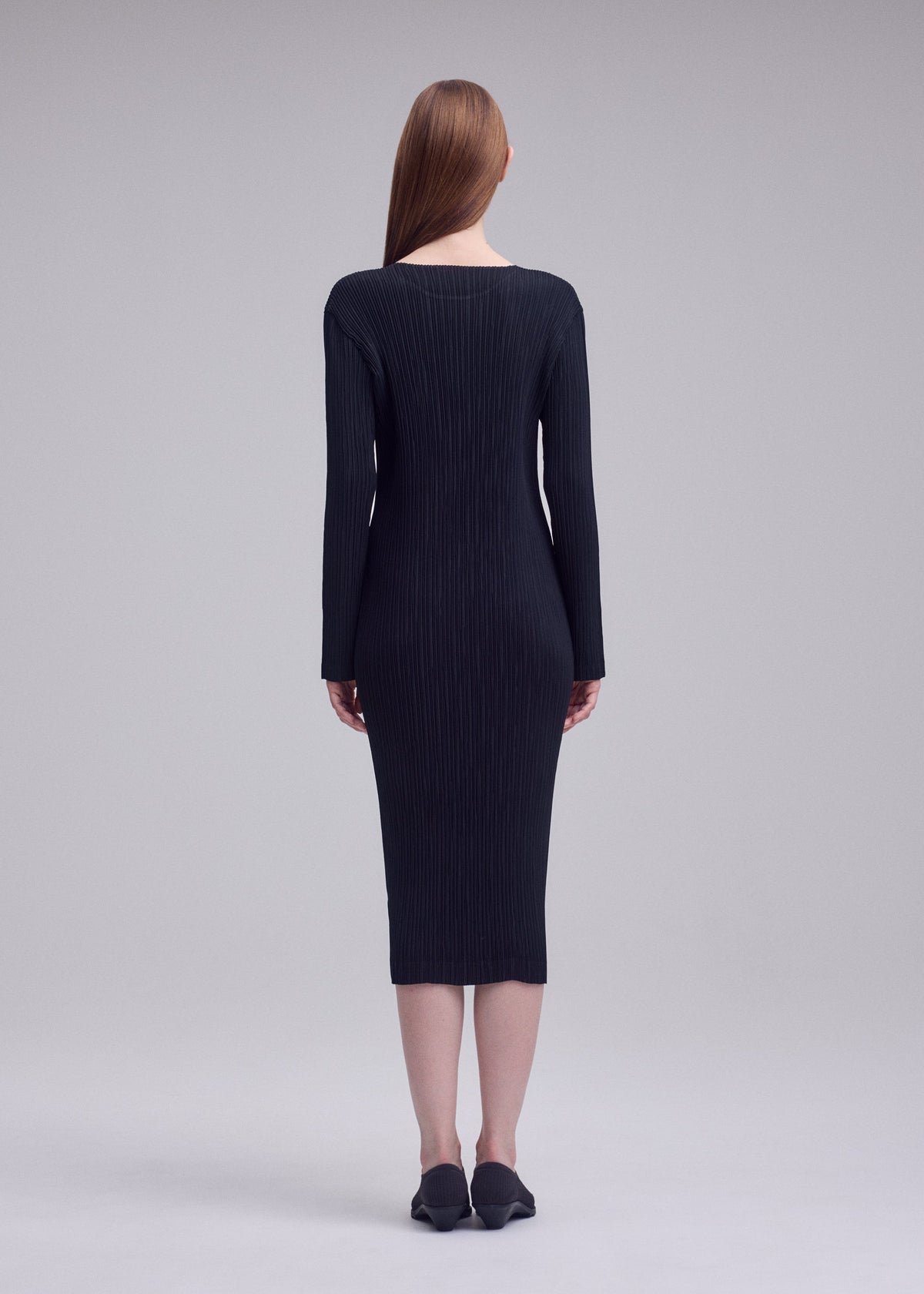 A woman with straight, long brown hair faces away, wearing the ISSEY MIYAKE HATCHING PLEATS fitted, long-sleeve black ribbed dress and black shoes against a plain gray background. view 5 of 5