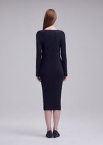 A woman with straight, long brown hair faces away, wearing the ISSEY MIYAKE HATCHING PLEATS fitted, long-sleeve black ribbed dress and black shoes against a plain gray background.