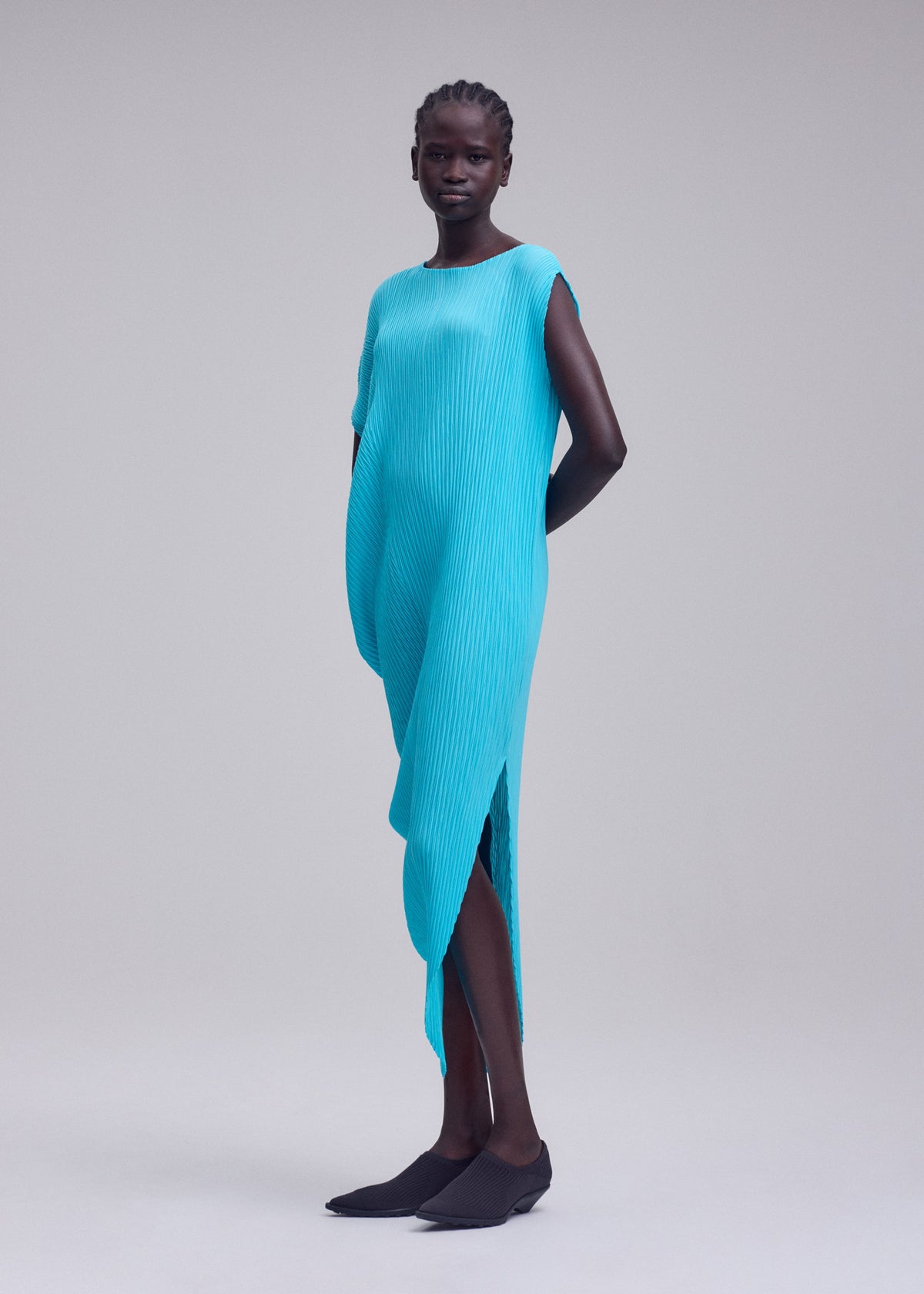 A woman stands in a studio wearing the LEAF PLEATS dress by ISSEY MIYAKE—a bright blue, asymmetrical, textured round-neck piece—paired with black flat shoes. Her hands are behind her back against a plain light gray background. view 6 of 7