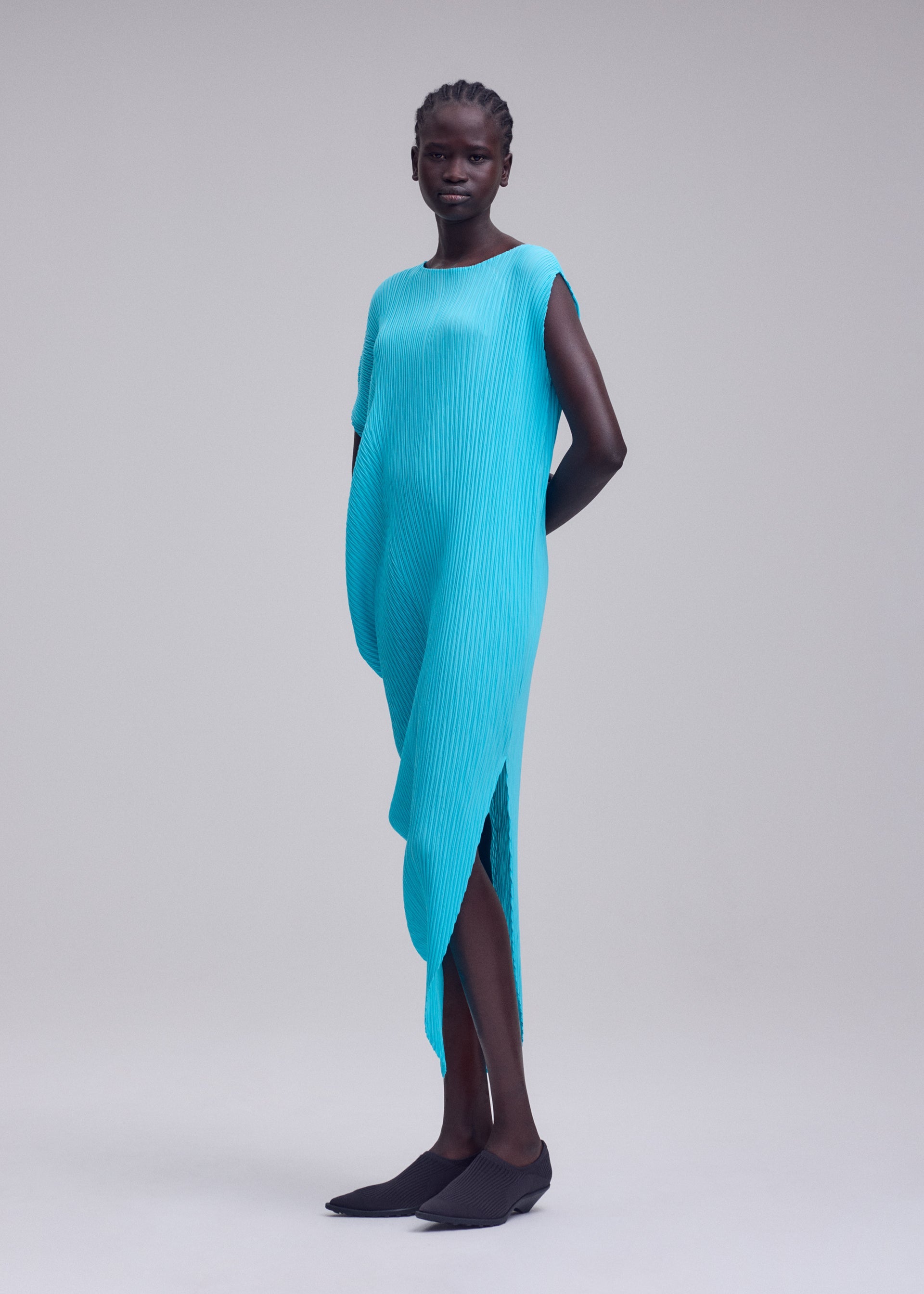 Zoomed view A woman stands in a studio wearing the LEAF PLEATS dress by ISSEY MIYAKE—a bright blue, asymmetrical, textured round-neck piece—paired with black flat shoes. Her hands are behind her back against a plain light gray background.