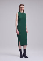 A woman poses in a studio wearing the ISSEY MIYAKE RHOMBUS PLEATS green midi dress with a side slit. The sleeveless design complements her straight brown hair, paired with black socks and shoes against a plain light gray background.