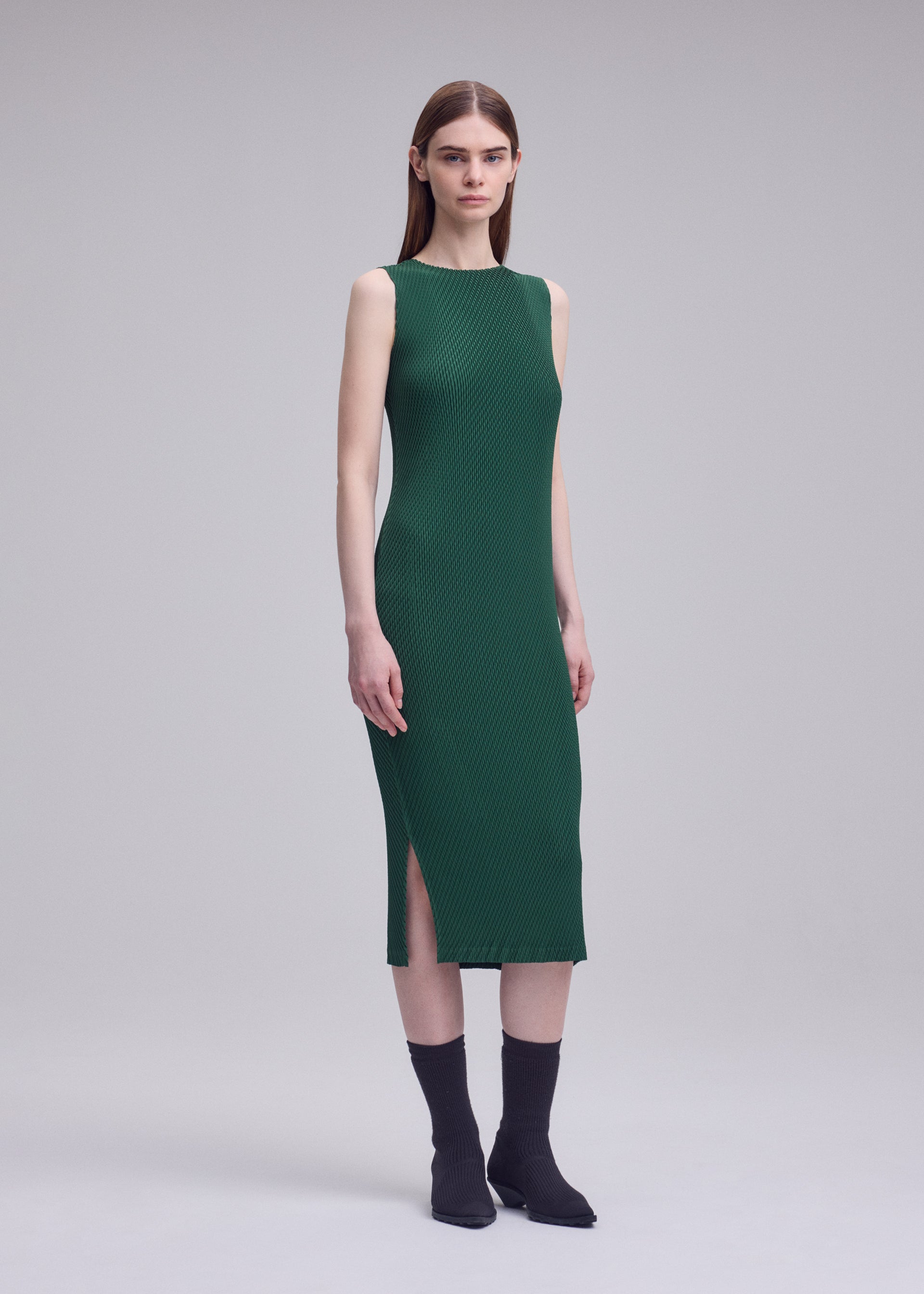 Zoomed view A woman poses in a studio wearing the ISSEY MIYAKE RHOMBUS PLEATS green midi dress with a side slit. The sleeveless design complements her straight brown hair, paired with black socks and shoes against a plain light gray background.