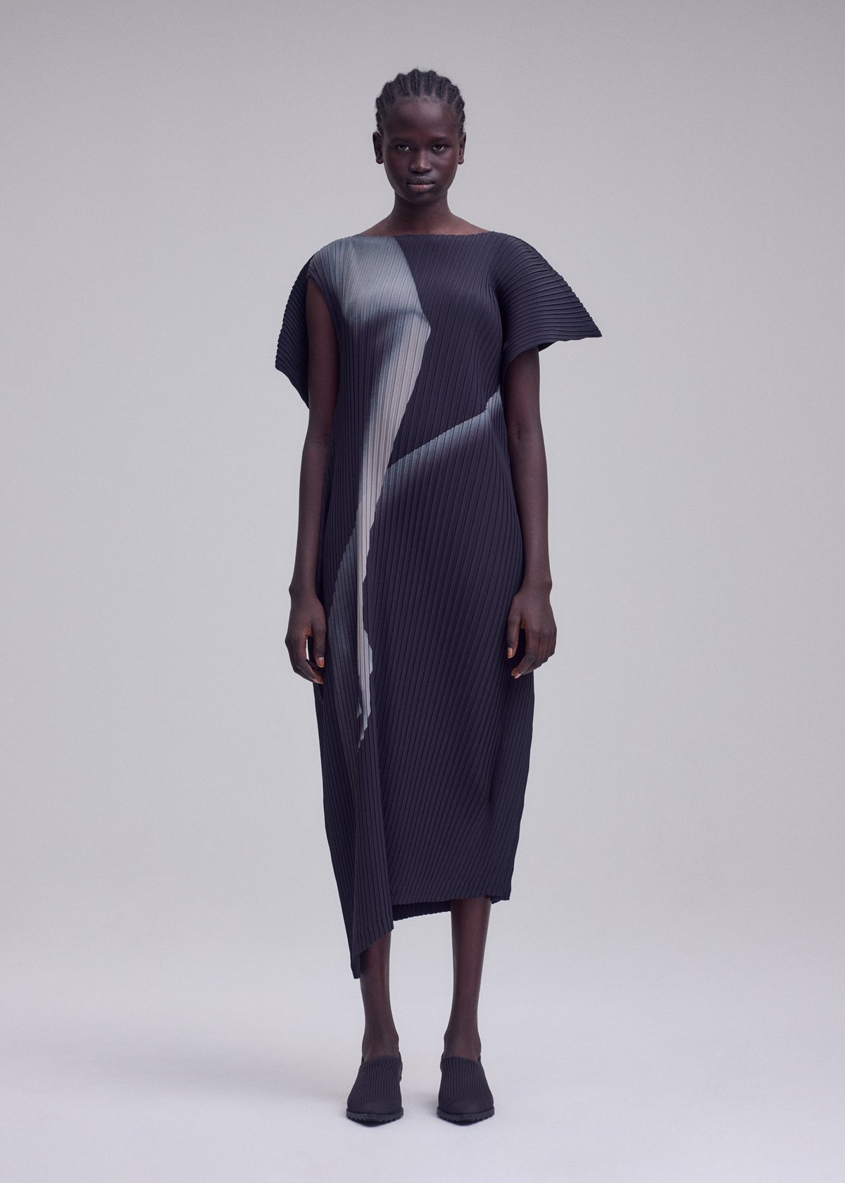 A person stands facing forward, wearing the ISSEY MIYAKE HANDS OF LOVER PLEATS black pleated midi dress, crafted from polyester crepe de chine and featuring abstract gray inkjet-printed patterns, paired with matching black shoes against a plain light gray background. view 4 of 5