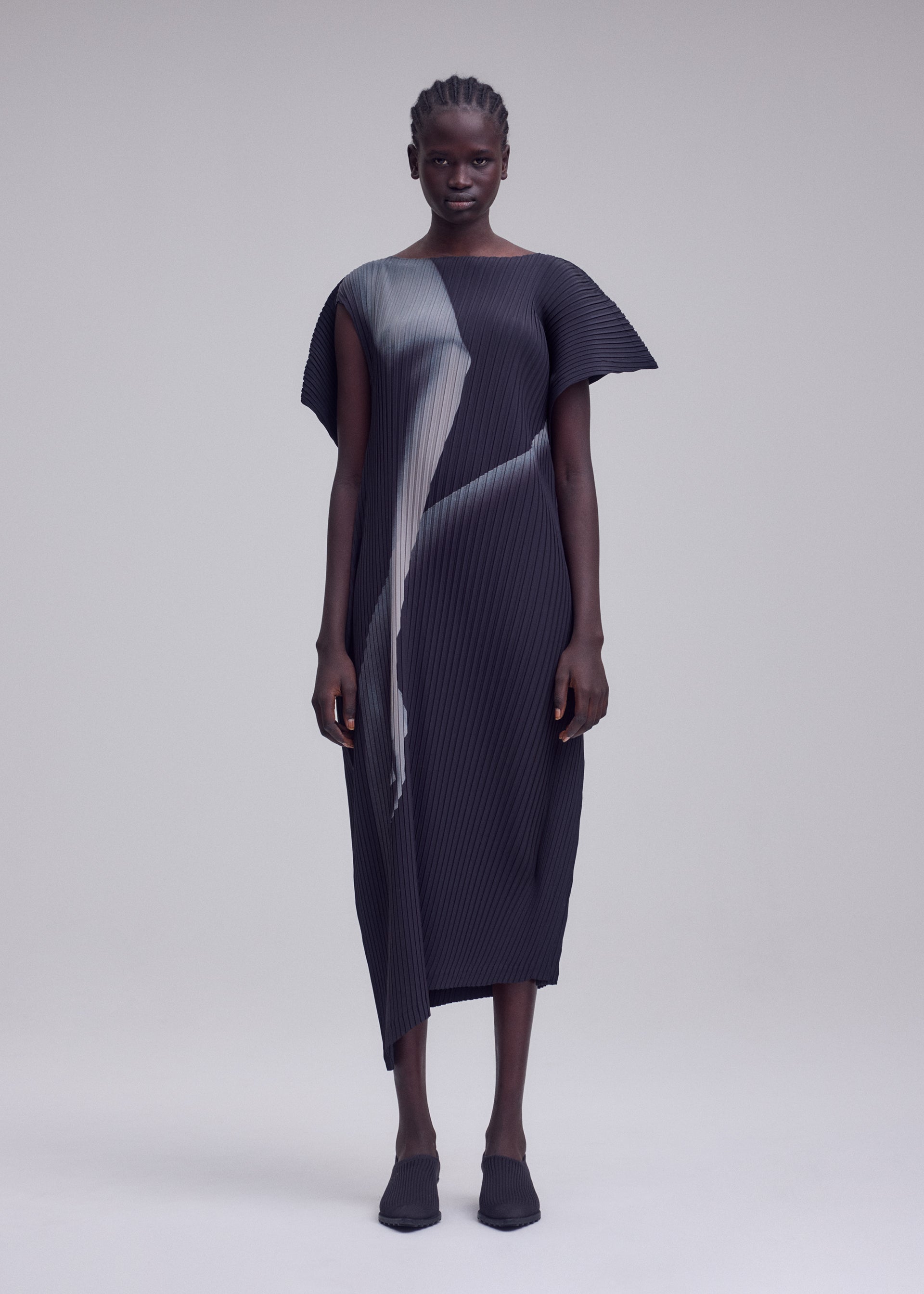 Zoomed view A person stands facing forward, wearing the ISSEY MIYAKE HANDS OF LOVER PLEATS black pleated midi dress, crafted from polyester crepe de chine and featuring abstract gray inkjet-printed patterns, paired with matching black shoes against a plain light gray background.