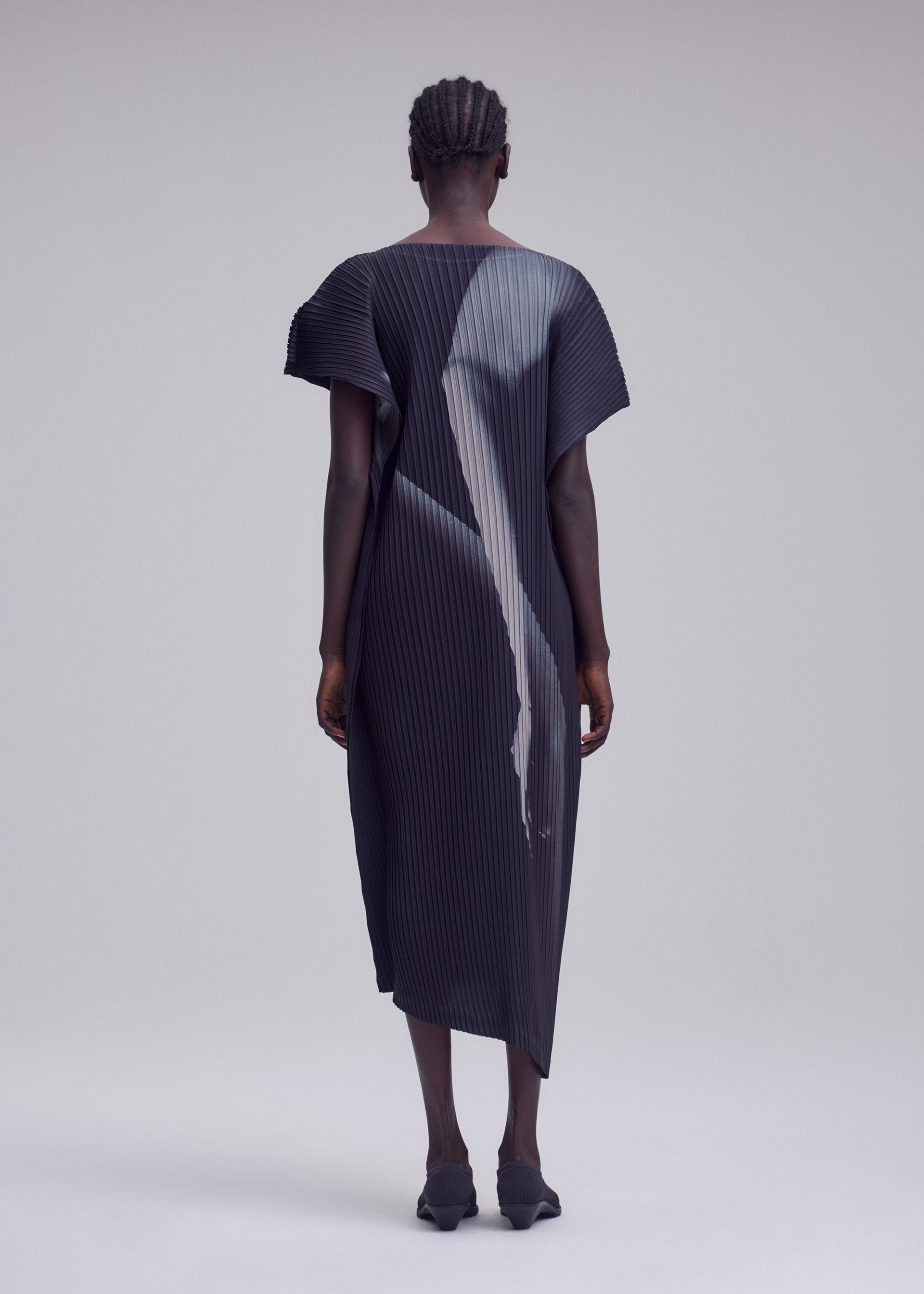 Zoomed view A person stands with their back to the camera, dressed in the HANDS OF LOVER PLEATS by ISSEY MIYAKE—a dark, short-sleeved dress made from polyester crepe de chine featuring original pleats. Its abstract design is vividly rendered using precise inkjet printing, set against a simple, light-colored background.