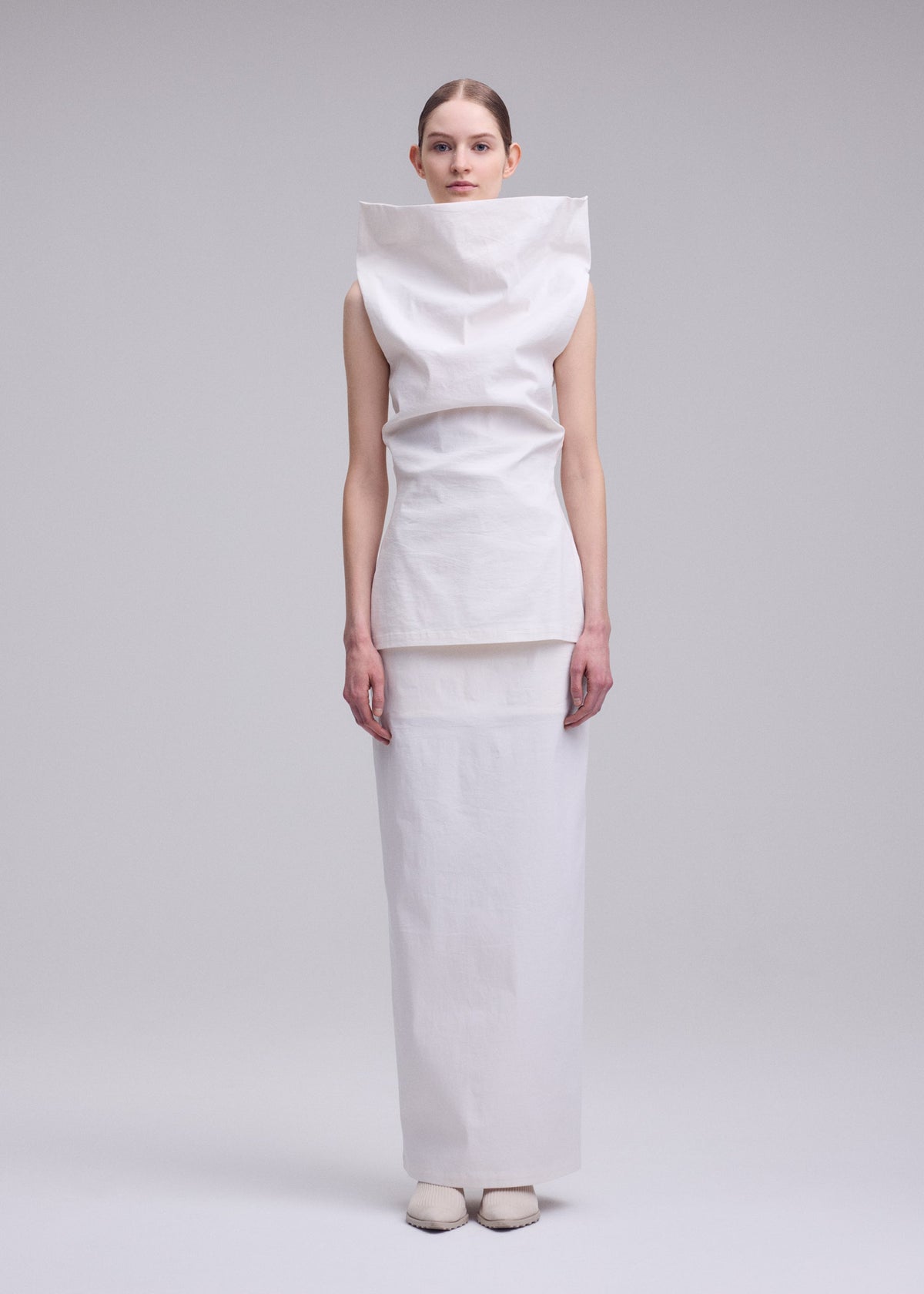 A person stands against a plain background, wearing the minimalist ALLOW high-neck sleeveless top and long skirt by ISSEY MIYAKE. Their hands rest by their sides, and they have a neutral expression. view 5 of 8