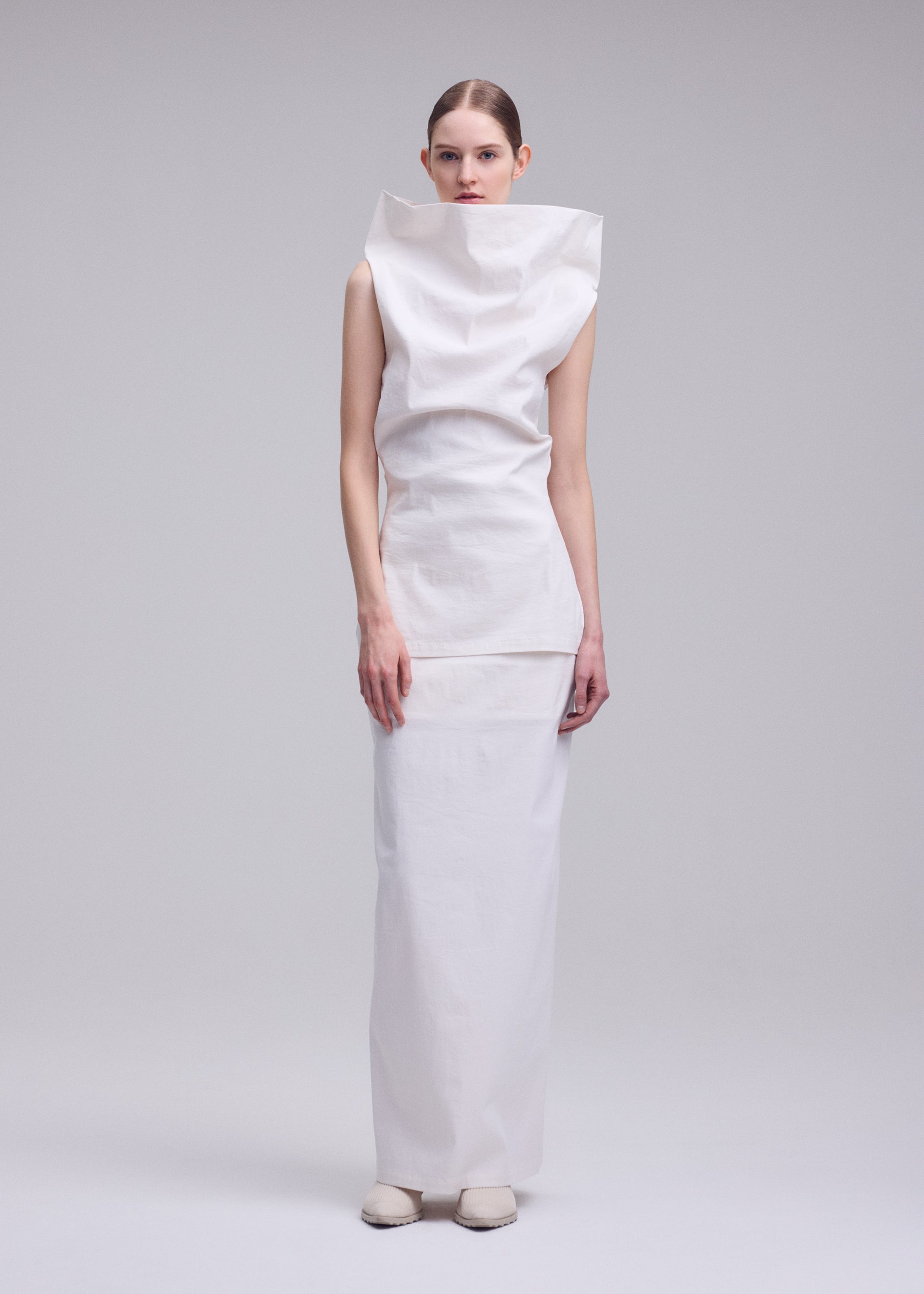 Zoomed view A person stands against a plain background in the ISSEY MIYAKE ALLOW dress, a unique sculptural white piece crafted from washi paper-blend with a high structured collar and long straight skirt, hair pulled back, looking directly at the camera.